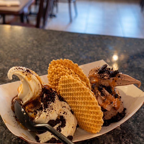 Two swirls of ice cream with caramel, cookie crumbles, and waffle crisps in a boat dish.