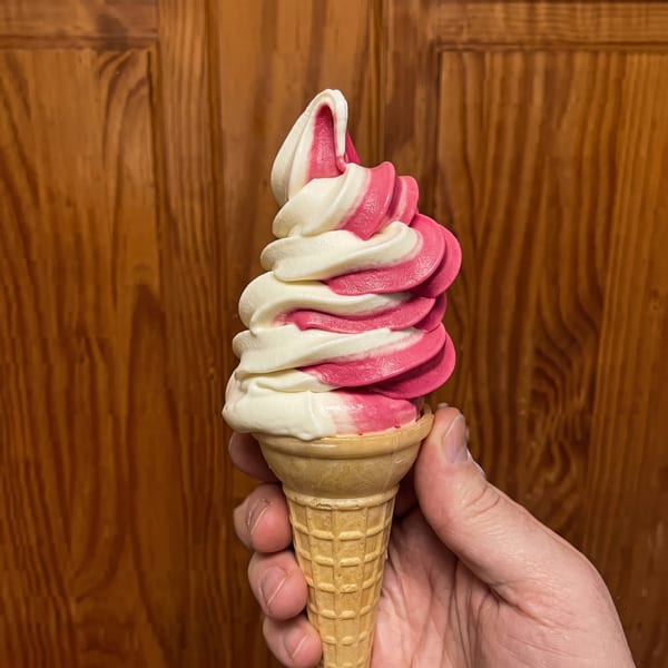 A hand holds a waffle cone with swirled pink and white soft-serve ice cream against a wooden door.