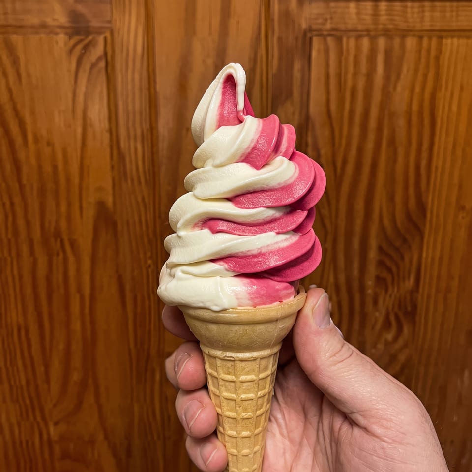 A hand holds a waffle cone with swirled pink and white soft-serve ice cream against a wooden door.